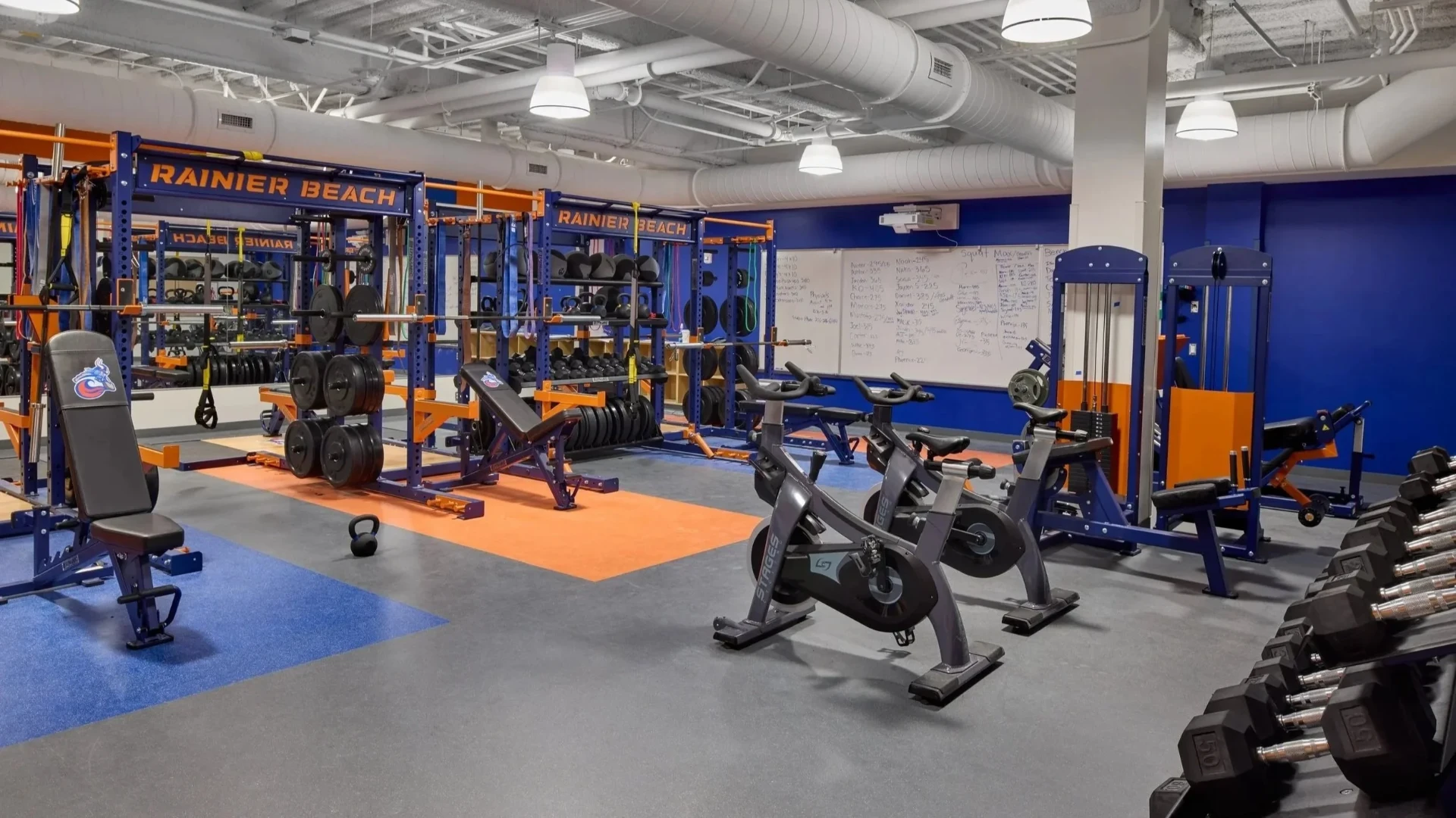 Rainier Beach weight room with blue and orange squat racks, benches and bikes